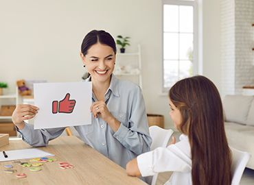 Happy proud female psychologist, therapist or teacher showing thumbs up paper card to little child. Kid receiving positive feedback from English language tutor. Therapy, learning, and success concept